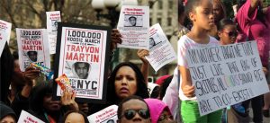 Two photographs depict people holding signs at a rally in protest of the death of Trayvon Martin. An African American woman in the photograph on the left holds a sign with the text 'one million hoodie march for Trayvon Martin,' in one hand, and a bag of skittles in the other. A young African American girl in the photograph on the right holds a sign with the text 'My mother taught me that just like that bag of skittles, all colors should be able to co-exist!!'
