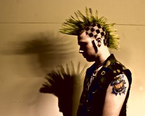 A young man with tattoos, a leather vest, and a spiky Mohawk haircut.