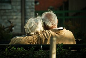 An elderly man and woman are shown from behind sitting on a bench. The man is shown wrapping his arm around the woman's shoulders
