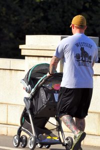 A photo of a man with many tattoos on his legs, pushing a baby stroller outside 