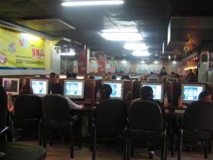 Many people sitting in chairs are shown staring at computer screens in a restaurant/cafe setting. Chinese posters can also be seen.