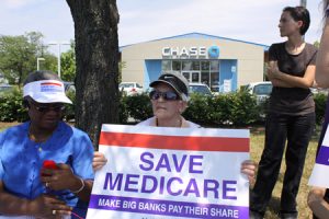 Two elderly women, one holding a red white and blue sign reading "Save Medicare: Make Big Banks Pay Their Share," are shown sitting under some trees and in front of a suburban bank building. A younger woman, dressed all in black, is shown behind and to the left of the other women.
