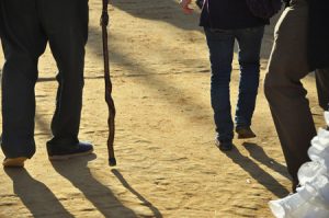 The legs of three men, one using a cane, are shown from behind walking on a dirt surface