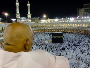 A man dressed in white is shown from behind looking down over the Kaaba, Islam's most sacred site. Hundreds of other people, dressed in all black or all white, can be seen circling a large black cube-like structure on the floor of a stadium-like structure.